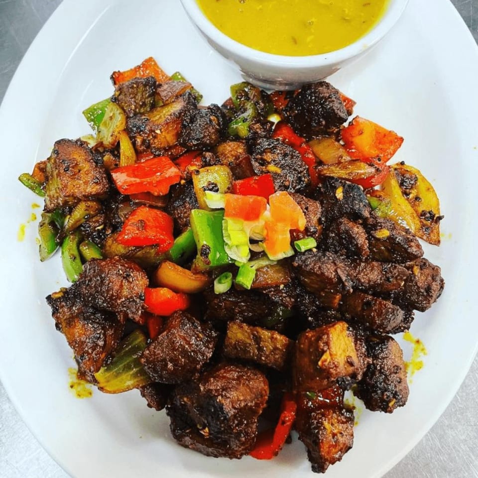 Indian dish with dark meat, colorful bell peppers, and a side of yellow sauce on a white plate.