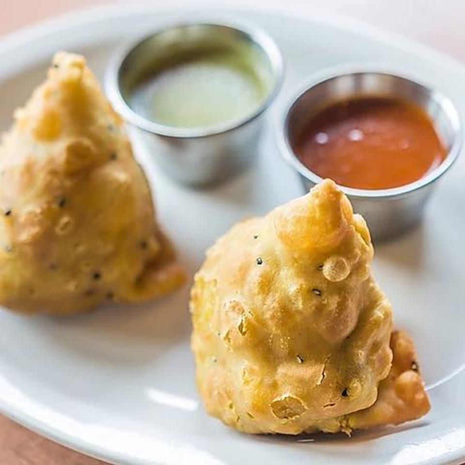Two golden-brown samosas with black seeds on a white plate, served with green and red sauces.